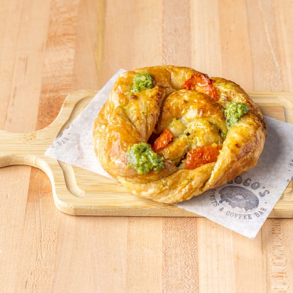 Golden twisted pastry with roasted tomatoes and pesto, served on a SOGO'S branded wooden board.
