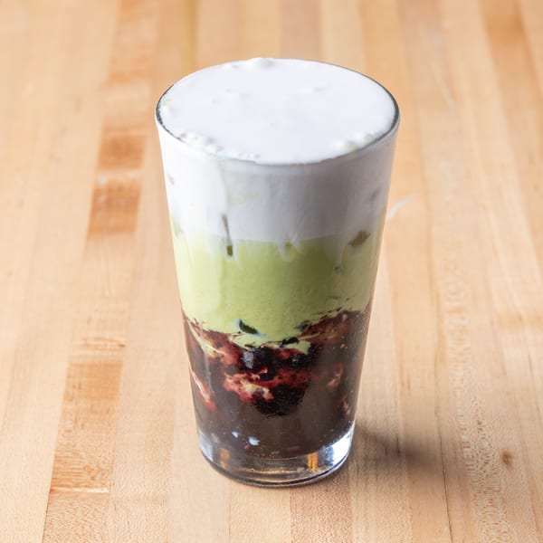Tall glass with layered white foam, green drink, and dark chunky bottom on wood.