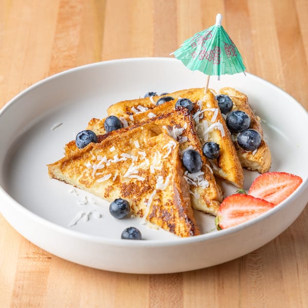 Golden French toast with blueberries, shredded coconut, strawberries, and a tiny green umbrella.