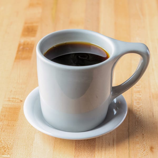 White mug of dark coffee on a saucer, resting on a light wooden table.