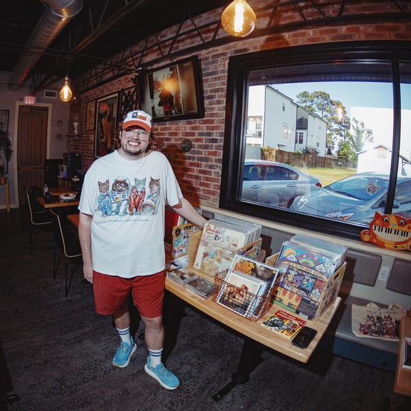 Man in a cat-themed shirt and hat stands next to a display of records and comics in a brick-walled room.
