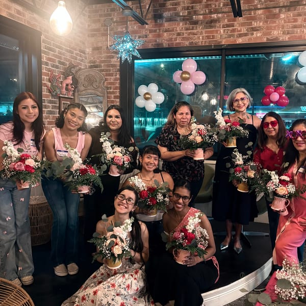 Group of women holding floral arrangements in a warmly lit room with brick walls and balloons.