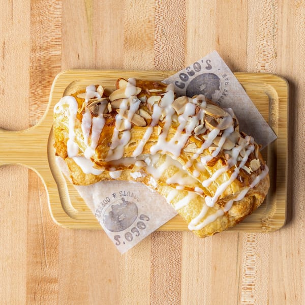 Flaky pastry with white icing and sliced almonds on a wooden board with Coco's branded paper.