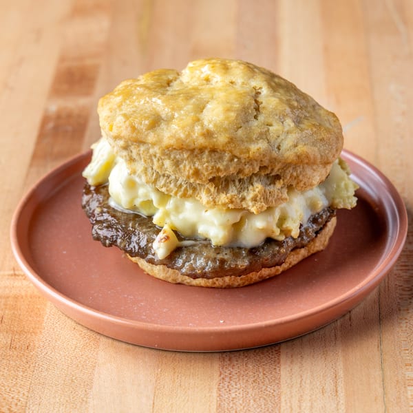 Golden biscuit sandwich filled with sausage and creamy scrambled eggs on a brown plate.